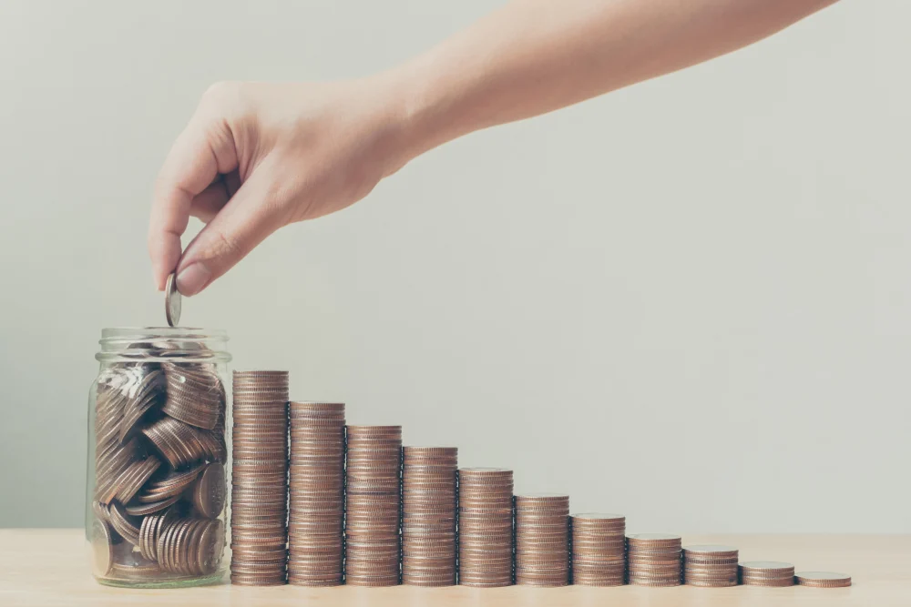 Glass mason jar filled with quarters, stacks of quarters leading up to it, woman hand putting quarters into jar