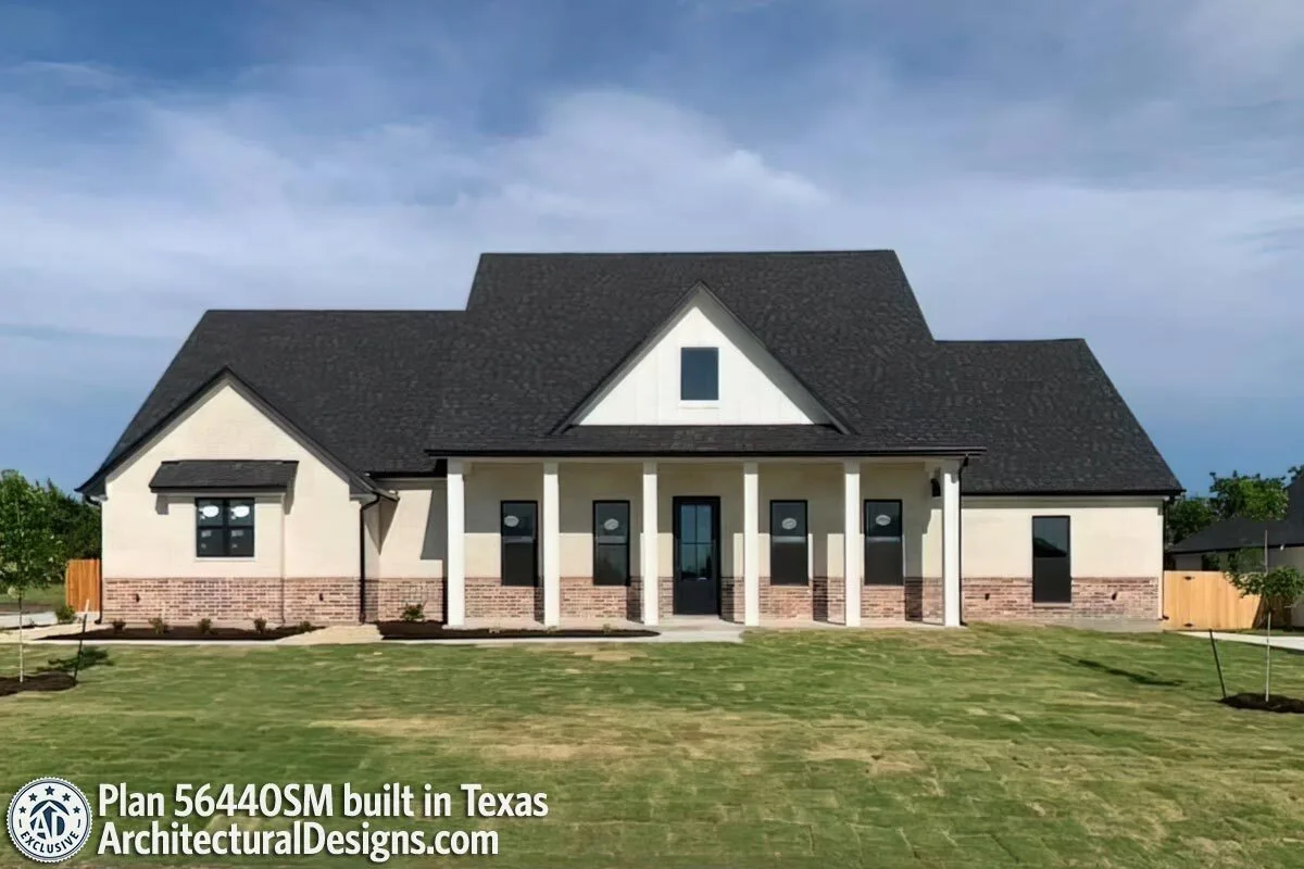 The front door of this contemporary farm home is a glass door, and the roof has a tone that is darker than the wall pillars and the white posts that are attached to the wall.